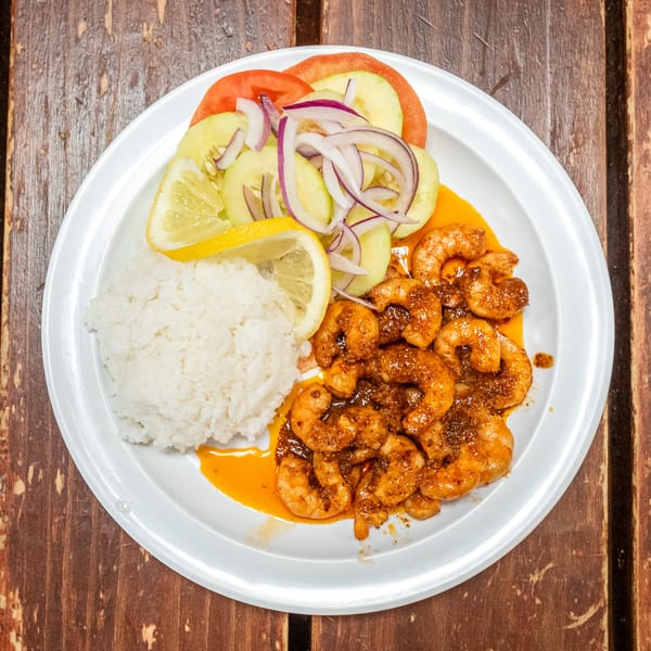 Spicy shrimp with white rice, sliced cucumbers, tomatoes, red onions, and lemon wedges on a white plate.