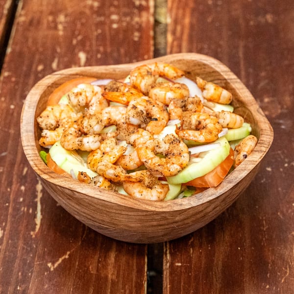 A wooden bowl filled with seasoned shrimp, sliced cucumbers, tomatoes, and onions on a rustic wooden table.