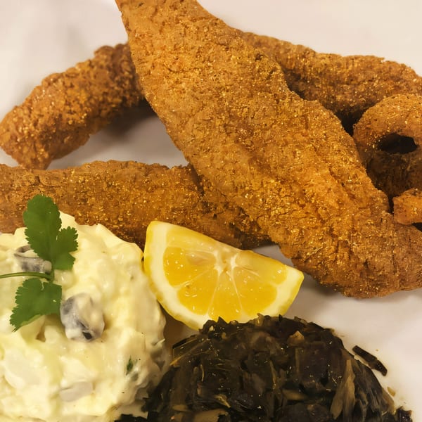 Fried fish fillets with potato salad, collard greens, and a lemon wedge on a white plate.